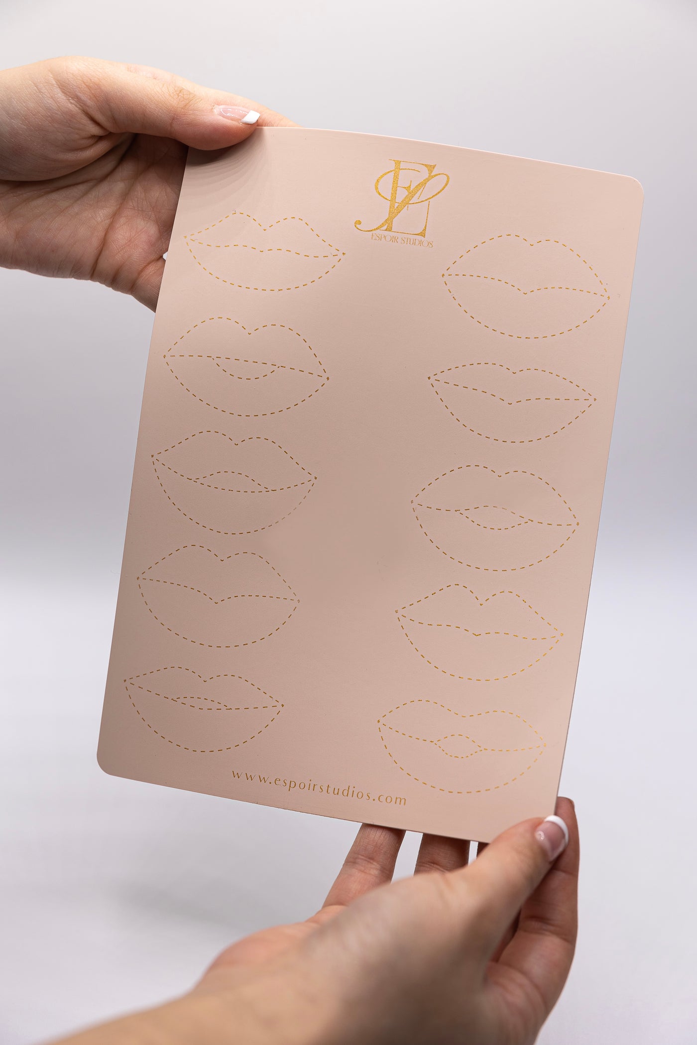 Two hands hold a pink practice sheet with outlines of lips, perfect for lip blushing practice. The sheet is from Espoir Studios, as indicated by the logo and website at the top and bottom.